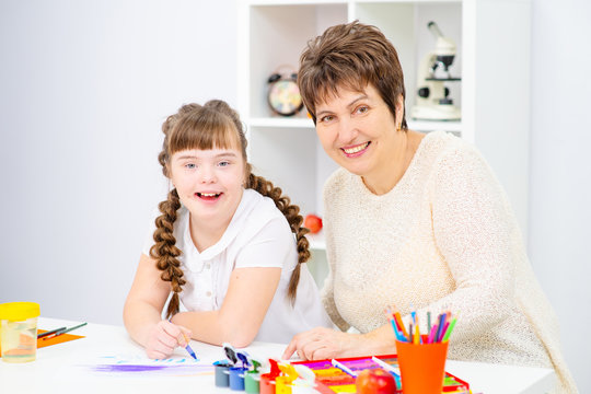 The girl and mother sitting at the table draw a picture at home. A girl with down syndrome at home with her mother is engaged in creativity