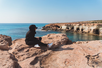 Woman with hoodie sitting alone on the cliff on asunny day. Cyprus, south coast