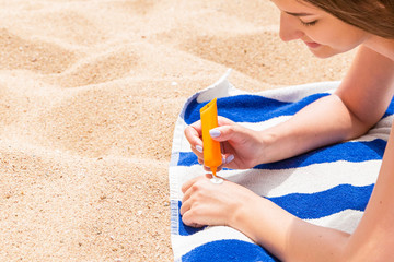 Attractive woman is lying on a striped towel at the beach and squeezing sunblock from a tube on her hand