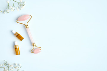 Jade roller for face and essential oil with flowers on blue background. Flat lay, top view, copy space. Rose quartz facial massage tool, beauty tool for skin care concept