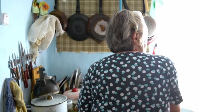 Grandma bakes pancakes in the cluttered kitchen. Outside the window the sun is shutting in 4K