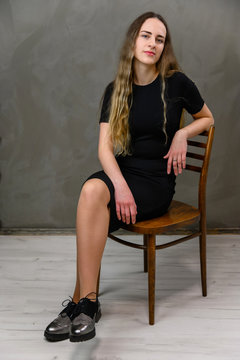 Vertical photo of a young pretty woman in a black dress on a gray background in the studio. A model with long hair is sitting on a chair.