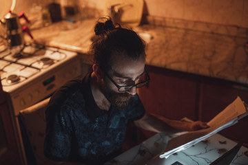 Stressed young hipster long hair bearded man is checking invoice in the kitchen.