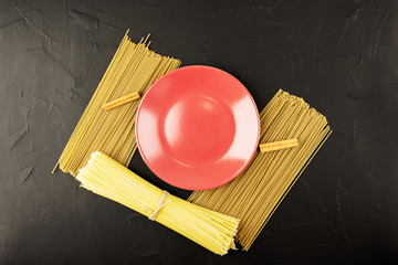 an empty red plate and bunches of different-colored spaghetti on a black textured background
