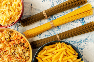 bundles of spaghetti of different colors and different paste in ceramic dishes on a white-blue textured background