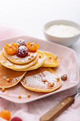 pancakes with raspberries, physalis, hazelnut and maple syrup on pink plate, sprinkled with powdered sugar, with fork and sour cream on pink kitchen towel