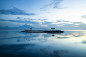 Sunrise seascape. Mountains and Agung volcano. Traditional gazebos on an artificial island in the ocean. Water reflection. Sanur beach, Bali, Indonesia.