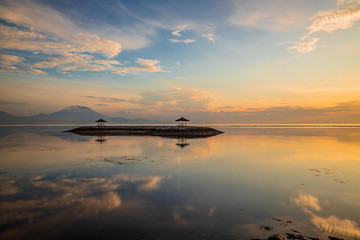 Sunrise seascape. Mountains and Agung volcano. Traditional gazebos on an artificial island in the ocean. Water reflection. Sanur beach, Bali, Indonesia.