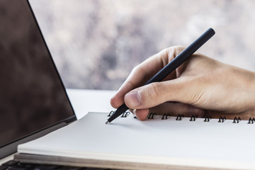 Man writes with a pen in notepad on laptop keyboard in a sunny office, business and education concept. Close up