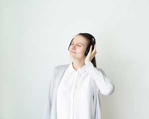 Young female is listening to music with headphones, girl standing in front of light gray wall