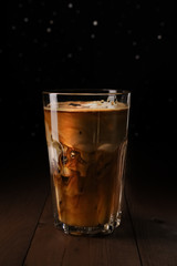 Ice latte coffee in tall glass on wooden table. Black coffee with milk in transparent glass. Black background with silver bokeh. Pouring cream into a glass with iced coffee.