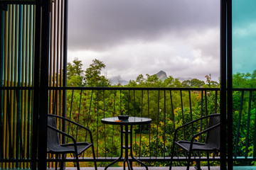 View from hotel room window with balcony to tropical forest during the storm with rain. Big shadow clouds and rainy weather. Two chairs on the side with simple table.