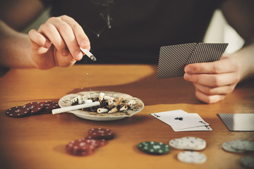 Home poker game. A man is sitting at a table holding cards in one hand and flicking the ash from...