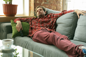 Obraz premium Watching TV lying on sofa. Caucasian man staying at home during quarantine because of coronavirus, COVID-19 spreading. Trying to spent time fun. Concept of healthcare and medicine, isolation.