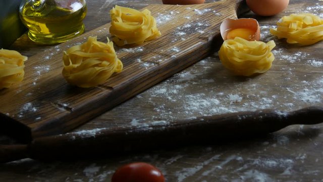 Tagliatelle or fettuccine pastas nests on a wooden kitchen board next to a broken egg yolk, cherry tomatoes, flour and olive oil in a rustic style. National italian food.