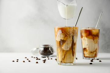 Milk is poured into coffee with ice on a light background. Two transparent glasses of refreshing iced coffee on a white table. Horizontal orientation, copy space.