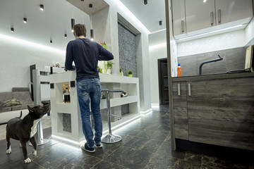 Young man cooking on modern kitchen. High tech interior in male style, gray concrete, dark wooden surfaces and black ceramic floor, designer light system. Black dog looks at owner. Indoors.