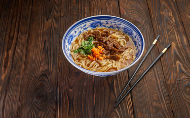 Chinese beef noodle soup in traditional blue bowl, fresh herbs and sliced carrot, pairs of chopsticks on dark wooden background and copy space. Asian food concept top view