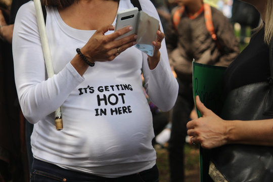 A pregnant woman at a climate change rally. She is holding her phone and wearing a white top reading “its getting hot in here” on the belly.
