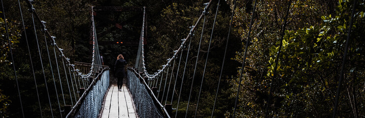 Hokitika Gorge, New Zealand, October 7, 2019: Stunning panorama of a blondie European woman crossing a suspension bridge surrounded by leafy trees