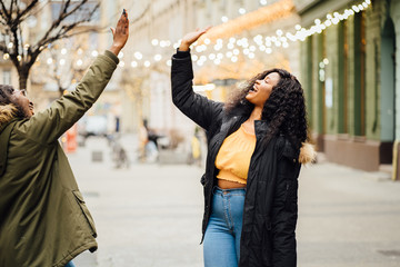 Happy exuberant young girl friends giving a high five slapping each others hand in congratulations as they sit together in a cafeteria enjoying a cup of hot coffee, multi ethnic viewed through glass