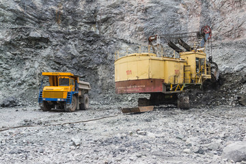 A daytime view of the iron ore mining quarry. In sunny weather. A heavy-duty heavy excavator loads ore onto a dump truck for transportation on an open iron ore mining career.