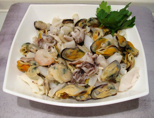 Sea cocktail: mussels, squids, small octopuses, shrimps with parsley leaves on a white square plate on a gray background