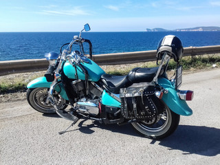 Fototapeta na wymiar Turquoise motorcycle parked by the sea with Capo Caccia on the background