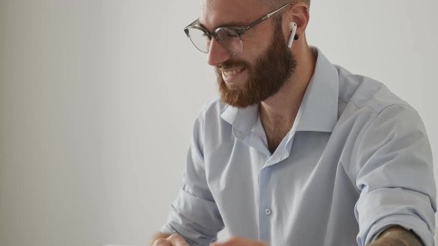 A confident bearded young man wearing wireless earphones and glasses is working with the documents while sitting at the table isolated over white wall