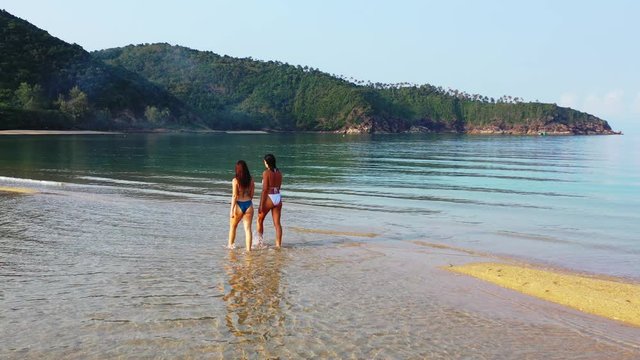 Two young women in swimsuits washing their feet on shallow lagoon with calm clear water near sandy exotic beach in Vietnam