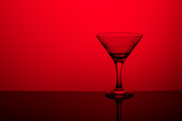 Glass goblet without wine on a thin leg stands on a mirror surface. Back light. Red background. Copy space.