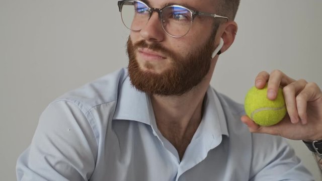 A positive confident young businessman wearing wireless earphones and eyeglasses is holding a tennis ball during his work over white wall