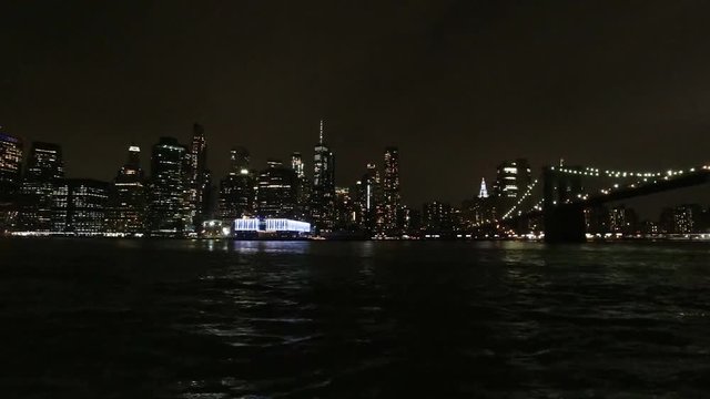 A timelapse of Manhattan skyline over the East River and Brooklyn Bridge at night with ferry passing by