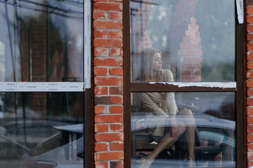 cute girl looks embarrassed at the floor while being outside the window of a brick building