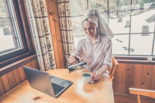 Young Woman Working From Home Office From The Cabin Or Cottage. Drinking Coffee. Working With Notebook. Woman With Towel On Her Head And Wearing Pyjamas Or Jammies