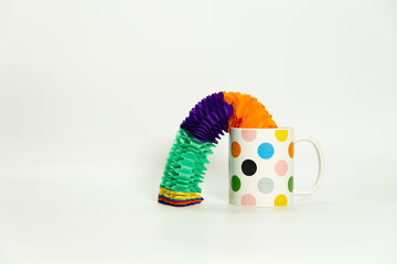Decorative image, on a white background a cup with colored dots and decorative elements coming out from inside or falling around. Garlands or and streamers of colors.