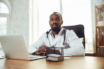 Doctor consulting for patient, working with laptop. African-american doctor during his work with patients, explaining recipes for drug. Daily hard work for health and lives saving during epidemic.