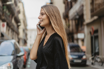 Girl crosses the road with cars and speak with her friend by the cellphone, she smiles and looks happy. Woman is using her gadget to communicate with her friends.