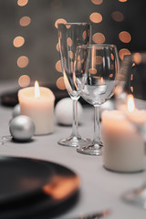 A table set in the restaurant. Serving a table. Grey artistic background. Black matte utensils, glass glasses. White candles and decor. Festive yellow lights garland in the background.