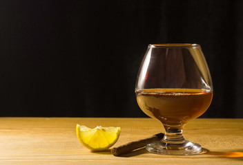 A glass glass of cognac on a wooden table, a slice of lemon and a cigar Free space for text on the side.