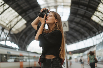 A young girl in a black shirt and black skirt with a backpack on her shoulder takes pictures of the railway station with her professional camera, there are few people at the station, she is smiling.