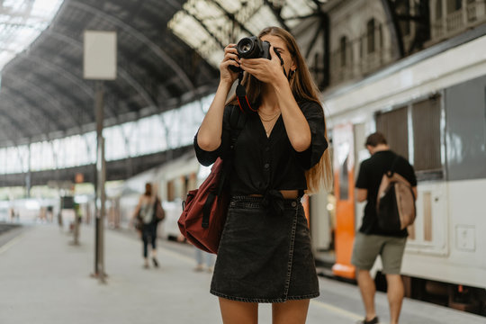 A young girl in a black shirt and black skirt with a backpack on her shoulder takes pictures of the station with her professional camera, concept of travelling and tourism and photography as a hobby.