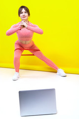 Athletic girl trains at home online watching videos on a laptop on a yellow background