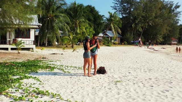Young women taking photographs holding coconuts on exotic beach with palm trees and white sand near resort cabins in Fiji