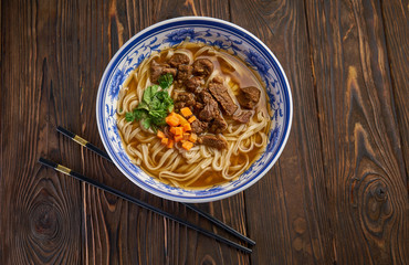 Chinese beef noodle soup in traditional blue bowl, fresh herbs and sliced carrot, pair of chopsticks on dark wooden background and copy space. Asian food concept top view
