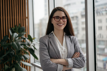 Portrait of young confident business woman with arms crossed standing in office. Successful manager in formal clothes and stylish eyeglasses looking at camera and smiling