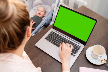 Mom is using a computer and spending time with her cute baby boy at home. Son and mummy working from home. Stay at home girl working remotely on laptop while taking care of her baby. Home office.