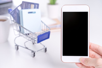 Woman holding smart mobile phone, concept of staying home online paying, electronic payment shopping on white table background with shop cart trolley, close up.