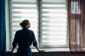 Woman's silhouette at window in the background light. Young woman in gown stands near window with blinds. Quarantine, self-isolation, stay home, self-preservation, coronavirus pandemic.