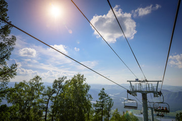 Ski lift in the mountains on sunny day against blue sky, white clouds, green hills and mountain lake. Mountain valley with cable car, view from top. Ski resort in summer.